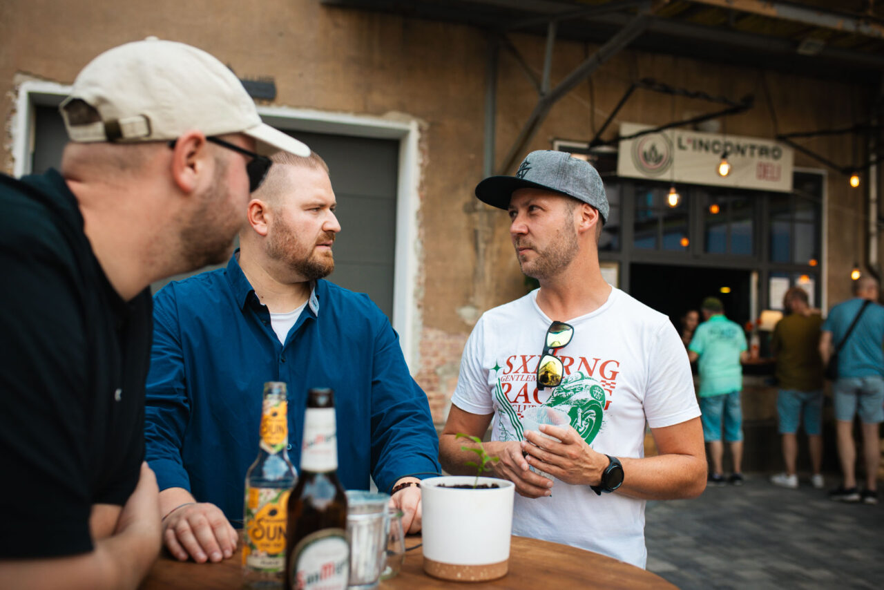 Business-Afterwork in entspannter Atmosphäre: Teilnehmer eines Events im Gespräch bei Kaltgetränken vor einer rustikalen Industrie-Location.