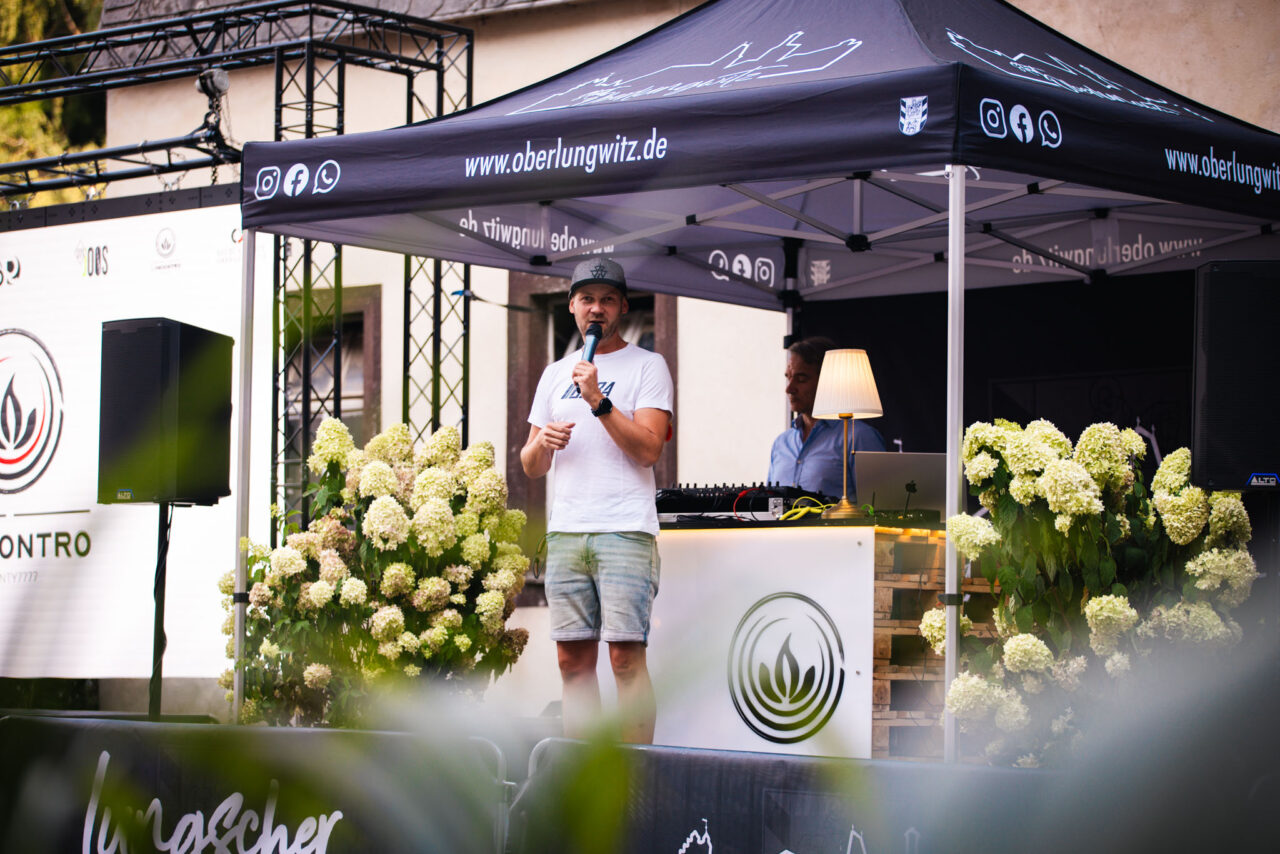 Sprecher bei einer Outdoor-Veranstaltung unter einem gebrandeten Faltpavillon mit Blumen-Deko und DJ-Equipment im Hintergrund.