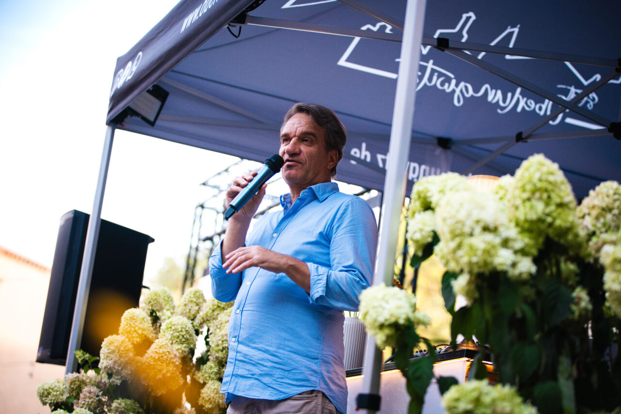 Sprecher bei einer Outdoor-Veranstaltung unter einem gebrandeten Faltpavillon mit Blumen-Deko und DJ-Equipment im Hintergrund.
