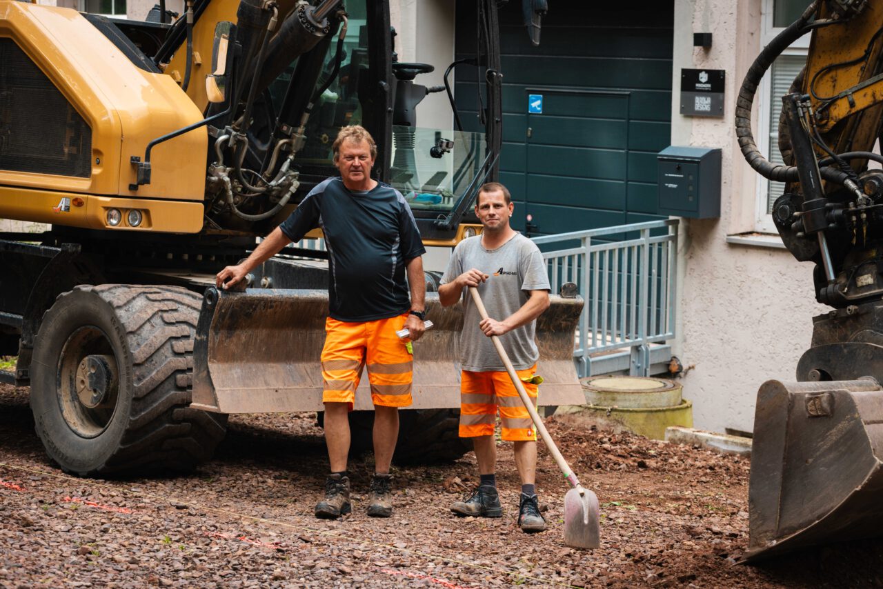 Professionelle Asphaltierung eines Gewerbehofs: Zwei Männer posieren bei Vorarbeiten unter Einsatz eines Baggers von Asphalt-Bau Chemnitz im Einsatz bei der Mindwork GmbH.